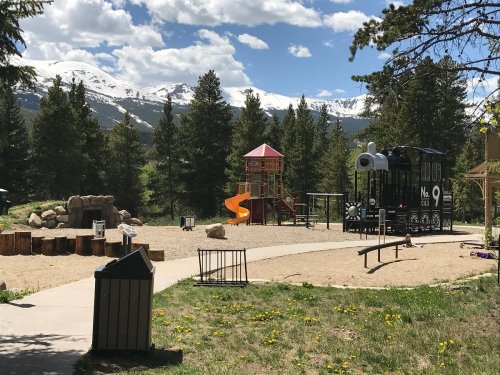 A playground with a slide and climbing structure, surrounded by trees and mountains under a blue sky with clouds.