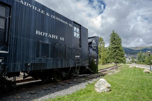 A vintage rotary snowplow train car on tracks, surrounded by grass and trees under a cloudy sky.