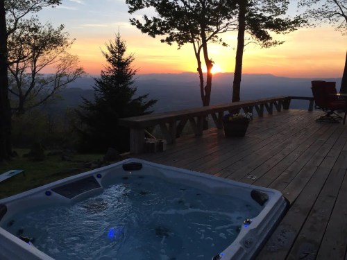 A hot tub on a wooden deck with a sunset view over mountains and trees in the background.