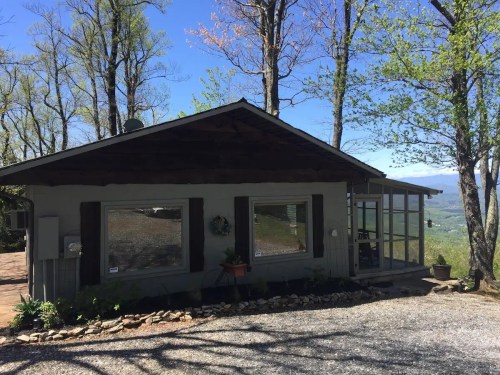 A cozy cabin surrounded by trees, featuring large windows and a gravel path, with a scenic view in the background.