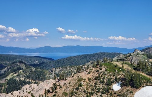 A panoramic view of a mountainous landscape with a lake in the distance under a clear blue sky with scattered clouds.