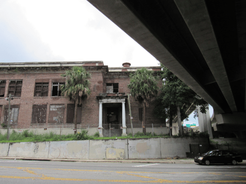 Abandoned brick building with boarded windows, palm trees, and an overpass above, set against a cloudy sky.