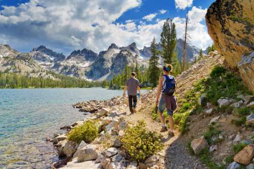 Two hikers walk along a rocky path by a clear lake, surrounded by mountains and lush greenery under a blue sky.