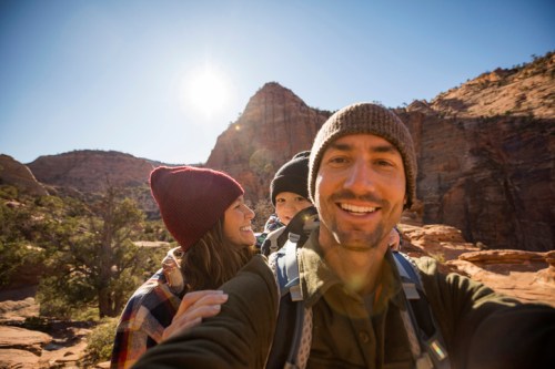 A smiling family poses for a selfie in a scenic outdoor setting with mountains and sunlight in the background.