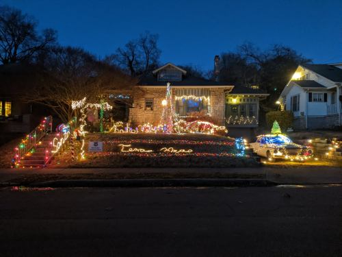 A festive house decorated with colorful lights, featuring a Christmas tree and a vintage car in the front yard.