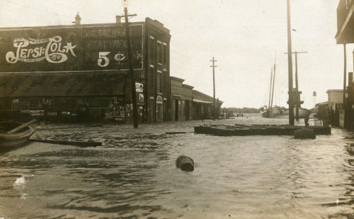 Historic photo of a flooded street with buildings and a Pepsi-Cola sign, showcasing high water levels and debris.