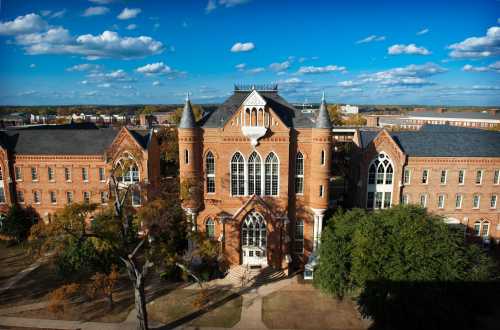 Historic brick building with towers, surrounded by trees and a blue sky with clouds.