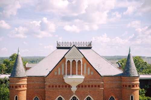 A brick building with a peaked roof and spires, surrounded by trees and a blue sky with fluffy clouds.