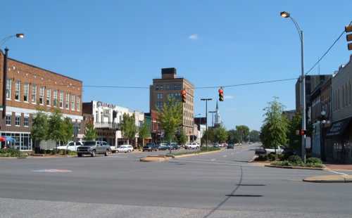 A sunny street view of a downtown area with shops, traffic lights, and trees lining the road.