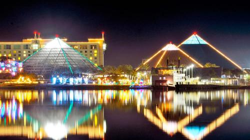 Colorful illuminated pyramids reflect in calm water at night, with a hotel and palm trees in the background.