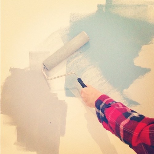 A hand holding a paint roller applies light blue paint to a wall, with a gray section already painted.