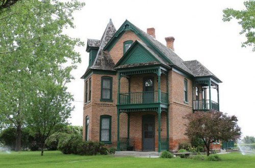 A charming Victorian-style brick house with green accents, surrounded by lush greenery and a well-maintained lawn.