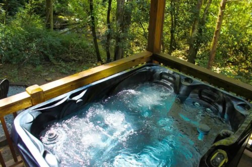A hot tub on a wooden deck surrounded by lush greenery and trees. Water bubbles gently in the tub.