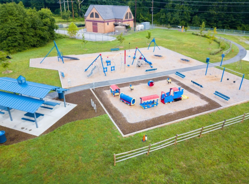 Aerial view of a playground with various equipment, picnic area, and grassy space surrounded by trees.