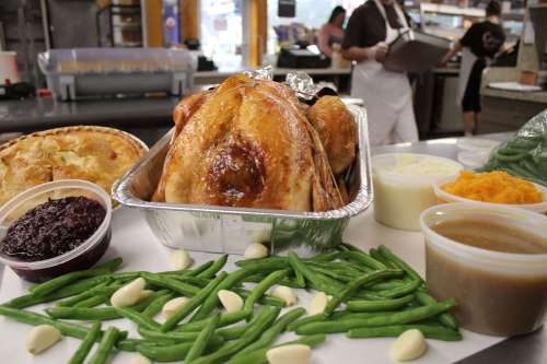 A roasted turkey in a tray surrounded by green beans, garlic, cranberry sauce, gravy, and a pie in a deli setting.