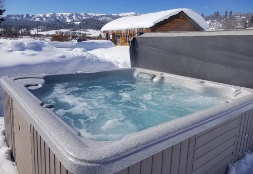 A hot tub filled with water sits in a snowy landscape, with mountains and a cabin in the background.
