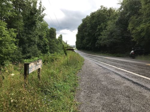 A gravel road lined with trees, featuring a sign for "Memorial Trail" and a distant view of a bridge.