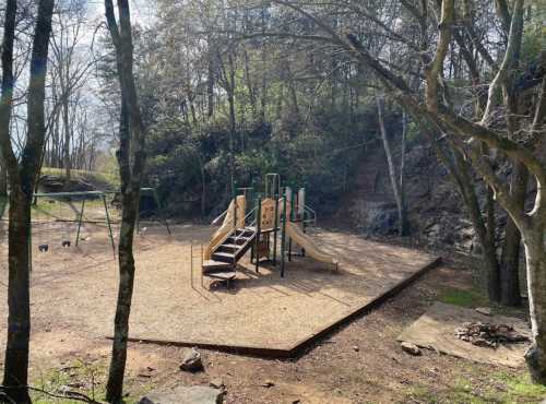 A playground with slides and swings, surrounded by trees and rocky terrain, on a sunny day.