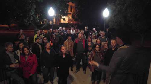 A crowd of people gathered outdoors at night, listening to a speaker near lampposts.