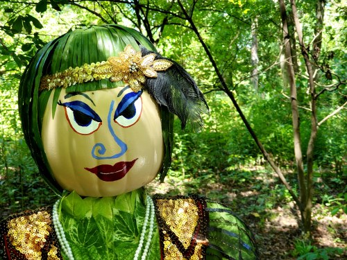 A whimsical scarecrow with a painted face, adorned in green leaves and sequins, stands amidst a lush forest backdrop.