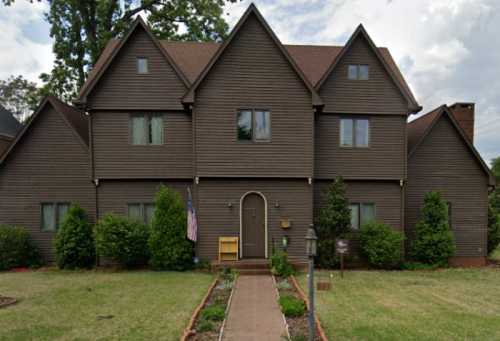 A large brown wooden house with multiple gables, surrounded by greenery and a pathway leading to the entrance.