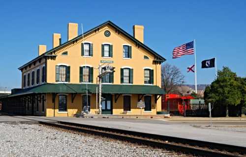 Historic yellow train station with American and state flags, set against a clear blue sky and railway tracks.