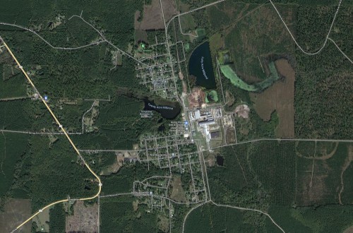 Aerial view of a small town surrounded by dense forest and water bodies, with roads and buildings visible.