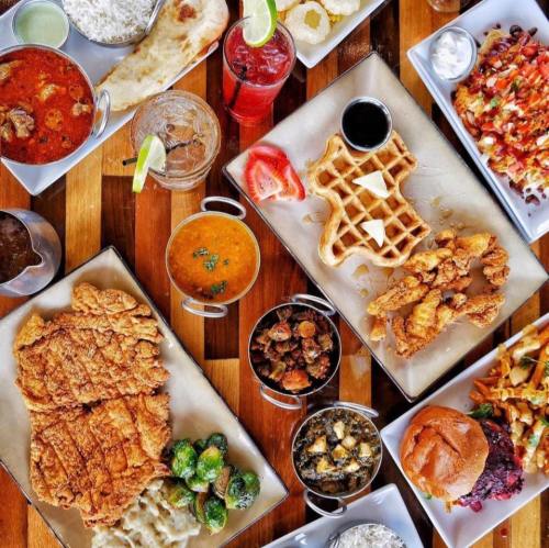 A vibrant spread of various dishes, including fried chicken, waffles, curry, and sides, arranged on wooden tables.