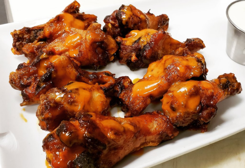 A plate of crispy chicken wings drizzled with spicy sauce, served with a small cup of dipping sauce.