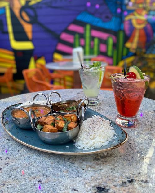 A colorful plate of Indian food with rice, two curries, and drinks on a table with a vibrant mural in the background.
