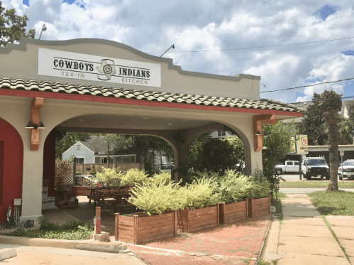 Exterior of Cowboys and Indians Kitchen, featuring a sign and landscaped planters under a cloudy sky.