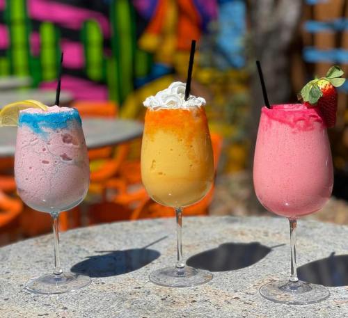Three colorful frozen drinks in tall glasses, topped with fruit and whipped cream, against a vibrant mural background.