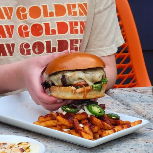 A person holds a burger topped with cheese and vegetables above a plate of fries drizzled with sauce.