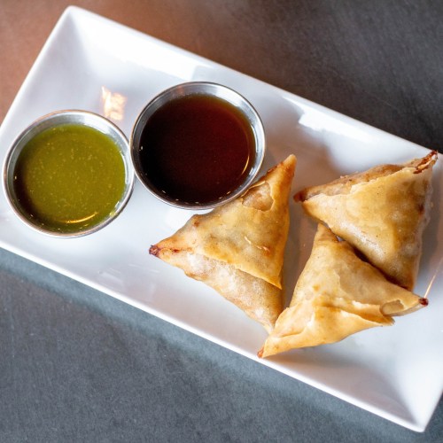Three golden-brown samosas on a white plate, accompanied by green and brown dipping sauces.