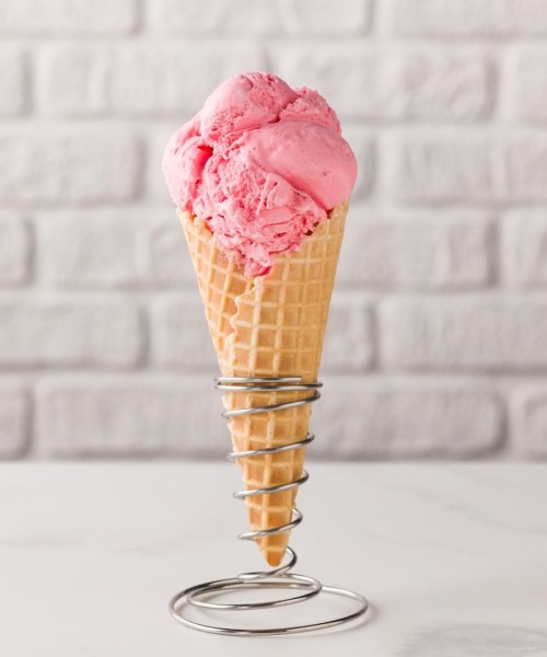 A pink ice cream cone with three scoops, placed in a metal holder against a textured white brick background.