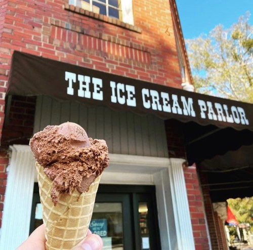 A hand holds a chocolate ice cream cone in front of an ice cream parlor with a brick facade.