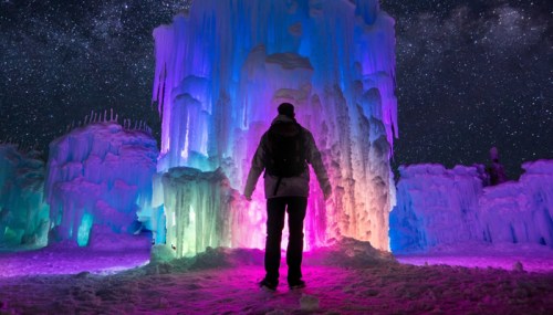 A person stands before a colorful ice formation under a starry night sky, illuminated in vibrant hues.