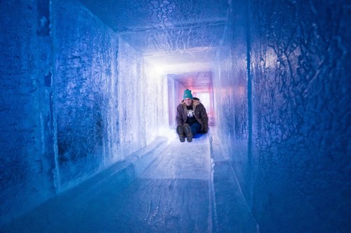 A person sits at the end of an icy tunnel, surrounded by blue ice walls, with soft light illuminating the space.