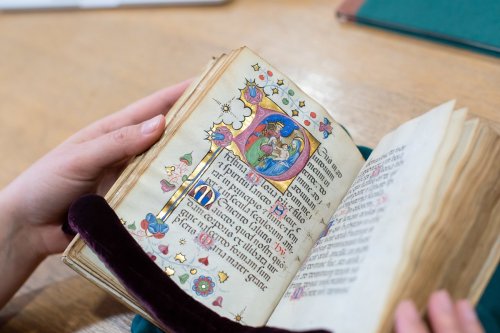 A person holds an open, illustrated medieval manuscript with colorful decorations and ornate text.
