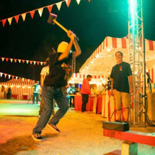 A person swings a hammer at a game booth, with colorful flags and tents in the background at a nighttime fair.