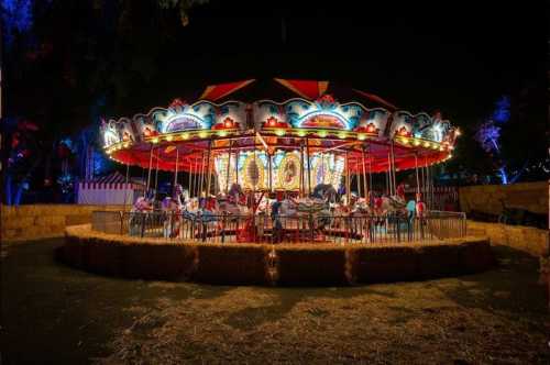 A brightly lit carousel at night, surrounded by hay bales and colorful lights, with people enjoying the ride.