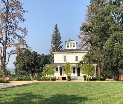 A large white house with a tower, surrounded by trees and a green lawn under a clear blue sky.