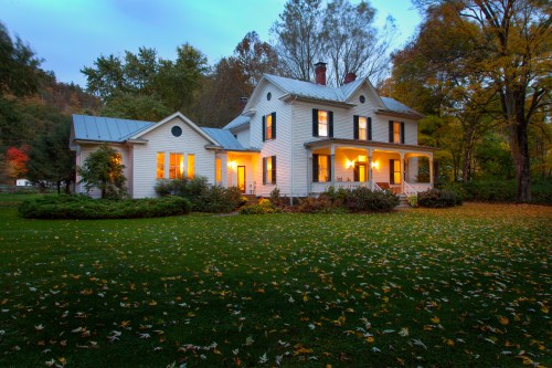 A charming white house with glowing windows, surrounded by autumn foliage and a lush green lawn at dusk.