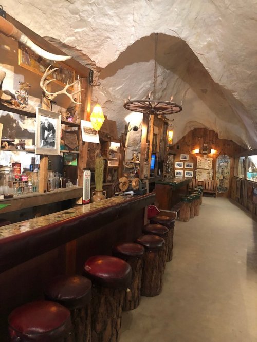 Interior of a rustic bar with wooden stools, stone walls, and decorative antlers, creating a cozy atmosphere.