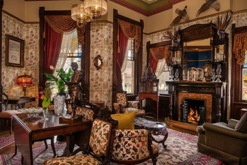 A cozy, elegantly decorated living room with vintage furniture, a fireplace, and ornate wallpaper.
