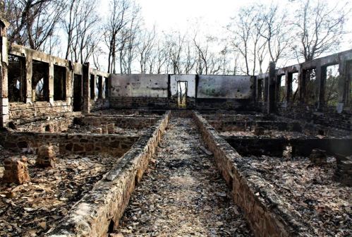 A charred, abandoned building with stone walls and remnants of a fire, surrounded by bare trees and debris.