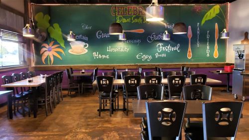 A cozy restaurant interior with wooden tables, black chairs, and a colorful mural featuring words like "family" and "friends."
