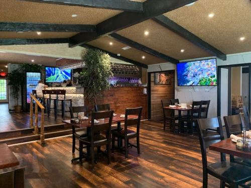 Interior of a restaurant featuring wooden floors, tables, a bar area, and large screens displaying images.