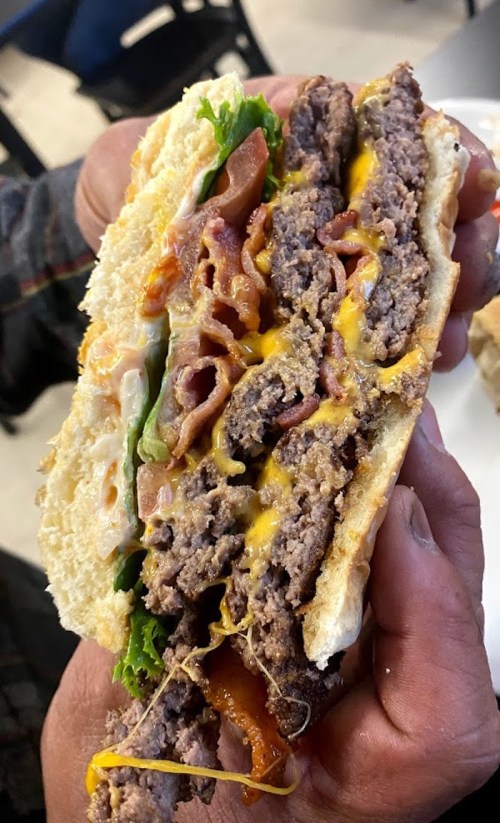 A close-up of a hand holding a thick, layered burger with beef patties, cheese, lettuce, tomato, and bacon.