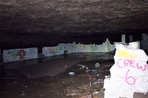A dimly lit underground space with graffiti on the walls and a wet floor, featuring crumbling stone structures.